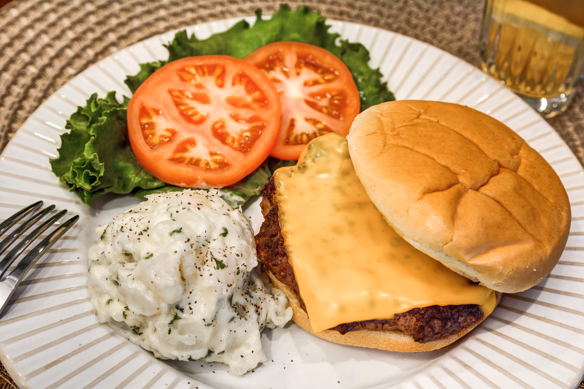 A plate with a cheeseburger featuring a slice of melted cheddar cheese on a beef patty inside a bun, two slices of tomato on a bed of lettuce, and a serving of mashed potatoes sprinkled with black pepper. A fork is placed on the left side of the plate and a glass of iced tea is visible in the background.
