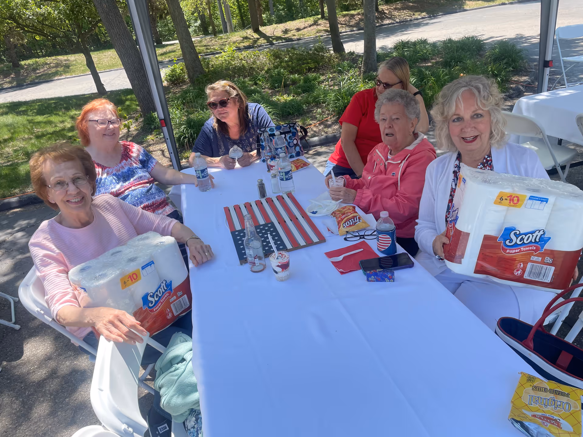 A group of five elderly women sitting around a white table outdoors under a canopy. Two women at the ends of the table are each holding a large pack of Scott paper towels. The table has an American flag decoration, water bottles, snacks, and cups with drinks. Trees and greenery are visible in the background.