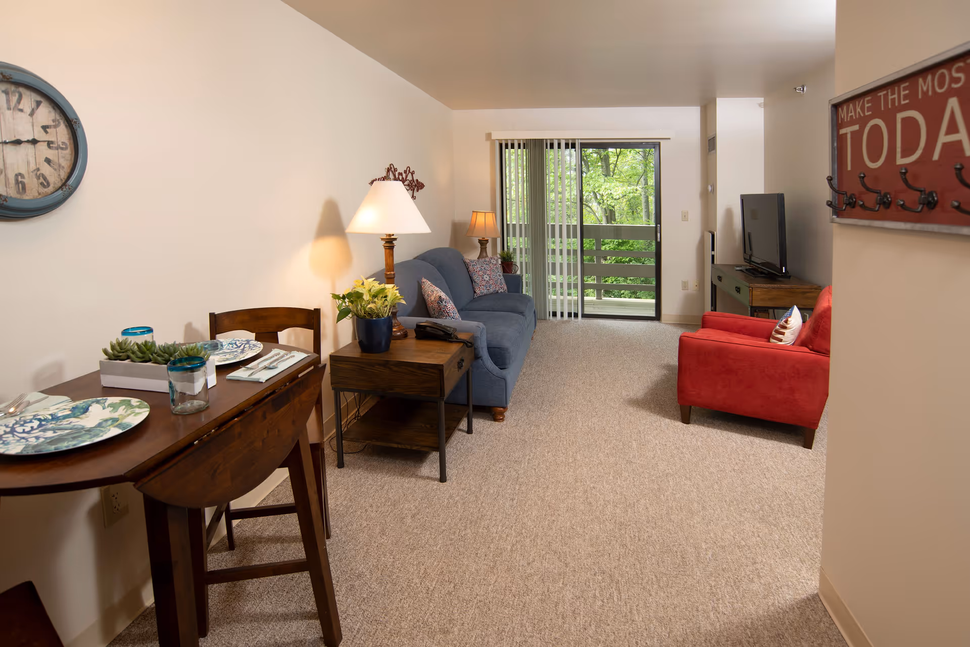 A cozy living room and dining area in a retirement community apartment. The room features a small wooden dining table set for two with plates and glasses, a blue sofa with patterned pillows, a red armchair, a wooden side table with a lamp and plant, a TV on a wooden stand, and a sliding glass door leading to a balcony with a view of green trees. A wall clock and a decorative sign with hooks are also visible.