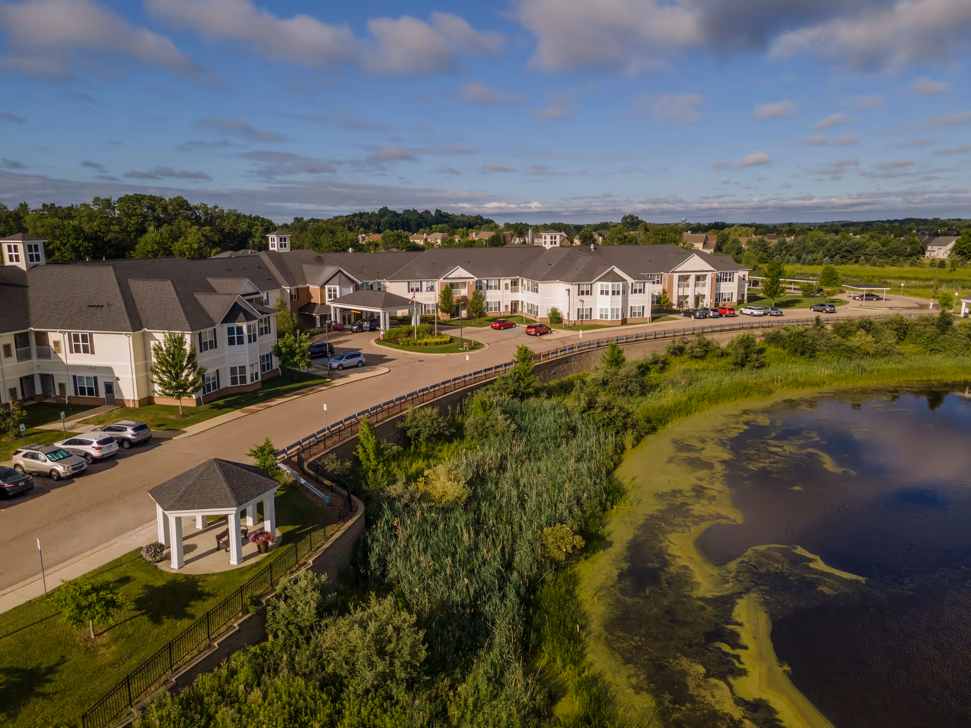 Aerial view of Olivia's Assisted Living facility showing a large, multi-wing building with a gray roof and white exterior. The building is surrounded by a parking lot with several cars, green lawns, trees, and a small white gazebo. In the foreground, there is a pond with green algae along the edges, and the sky above is partly cloudy with blue patches.