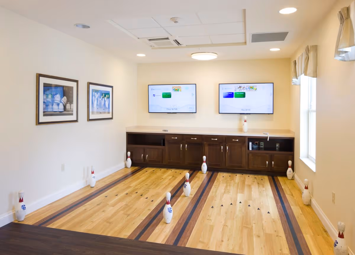 Indoor room with two small bowling lanes marked on a wooden floor, several bowling pins set up on the lanes and around the edges, two flat-screen TVs mounted on the wall above a wooden cabinet, two framed pictures of bowling pins on the left wall, and windows with curtains on the right side.