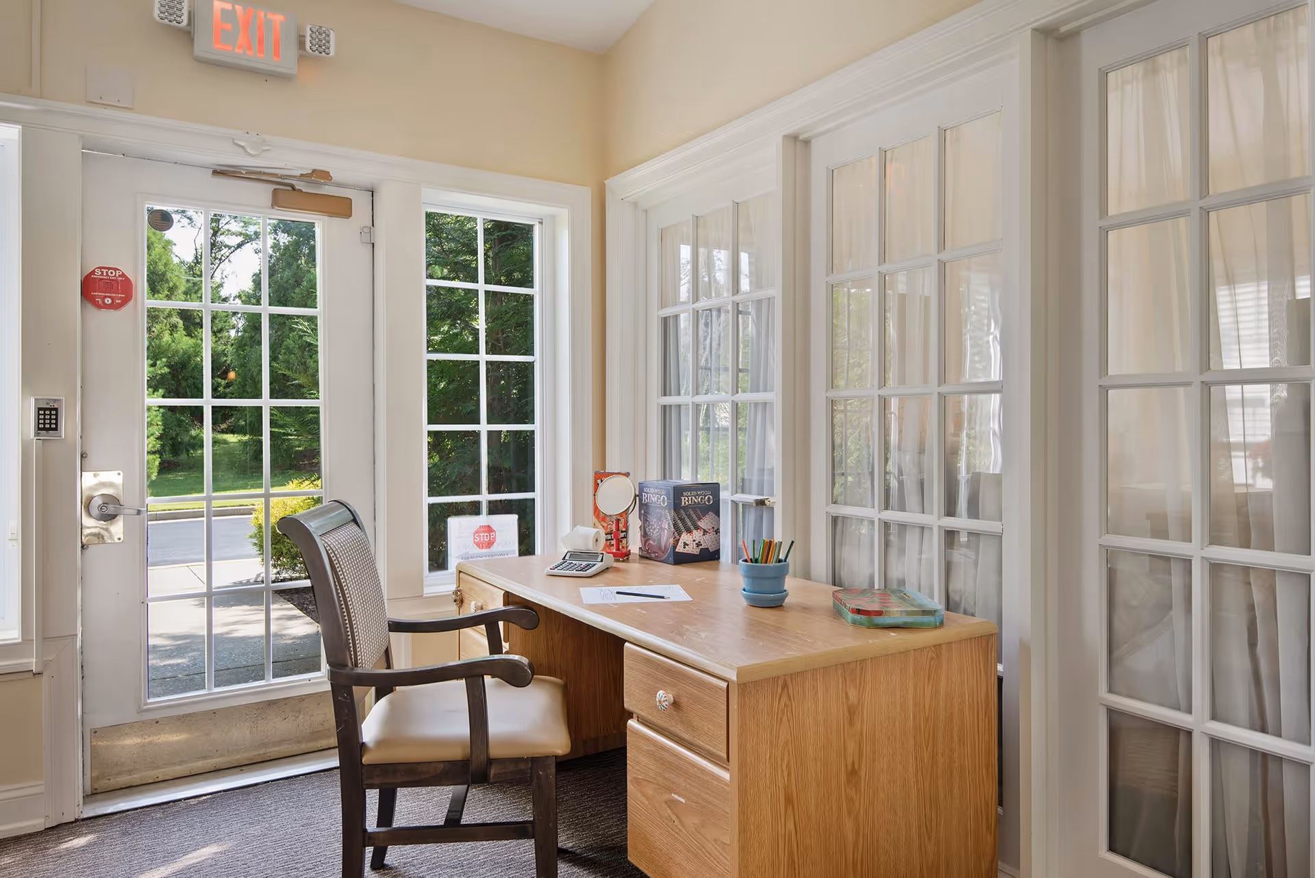 A small office area inside a senior living facility with a wooden desk and a chair. The desk holds a calculator, a bingo game box, a small mirror, a container with colored pencils, and some papers. The room has large glass-paned doors and windows letting in natural light, with a view of greenery outside. An exit sign is visible above the door.