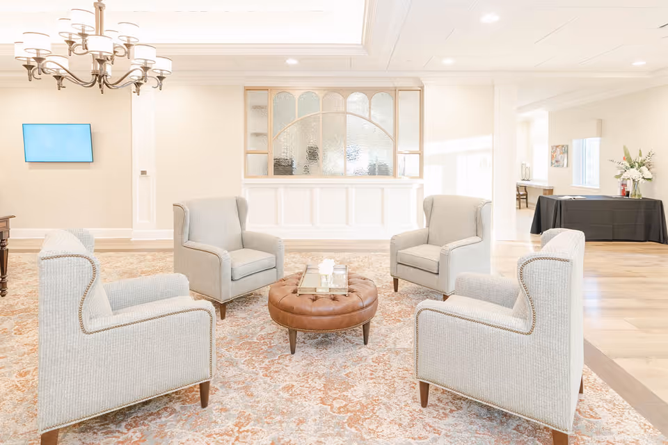 A bright and spacious sitting area with four light gray upholstered armchairs arranged around a round brown leather ottoman on a patterned area rug. The room features light-colored walls, a chandelier, a mounted flat-screen TV, and a decorative glass partition with wooden framing. In the background, there is a table covered with a black cloth and a vase of flowers.