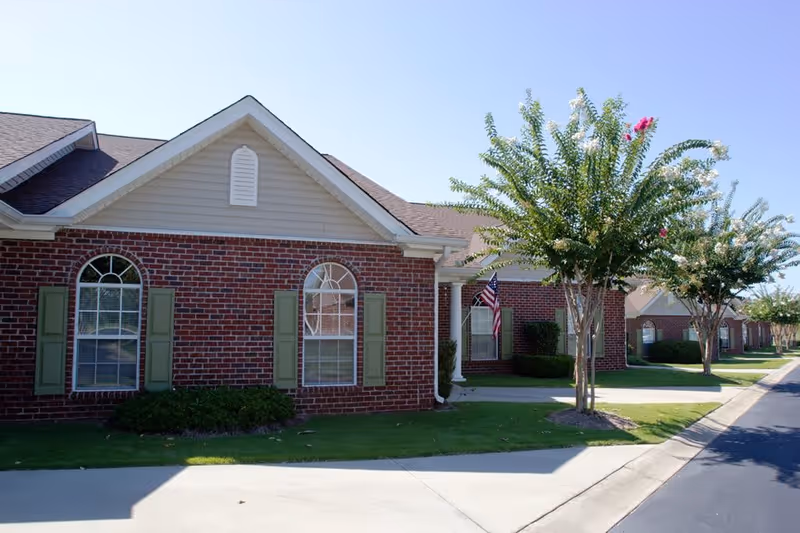 Exterior view of a single-story brick building with arched windows and green shutters, a small lawn with trees, and a sidewalk leading to the entrance. An American flag is displayed near the doorway.