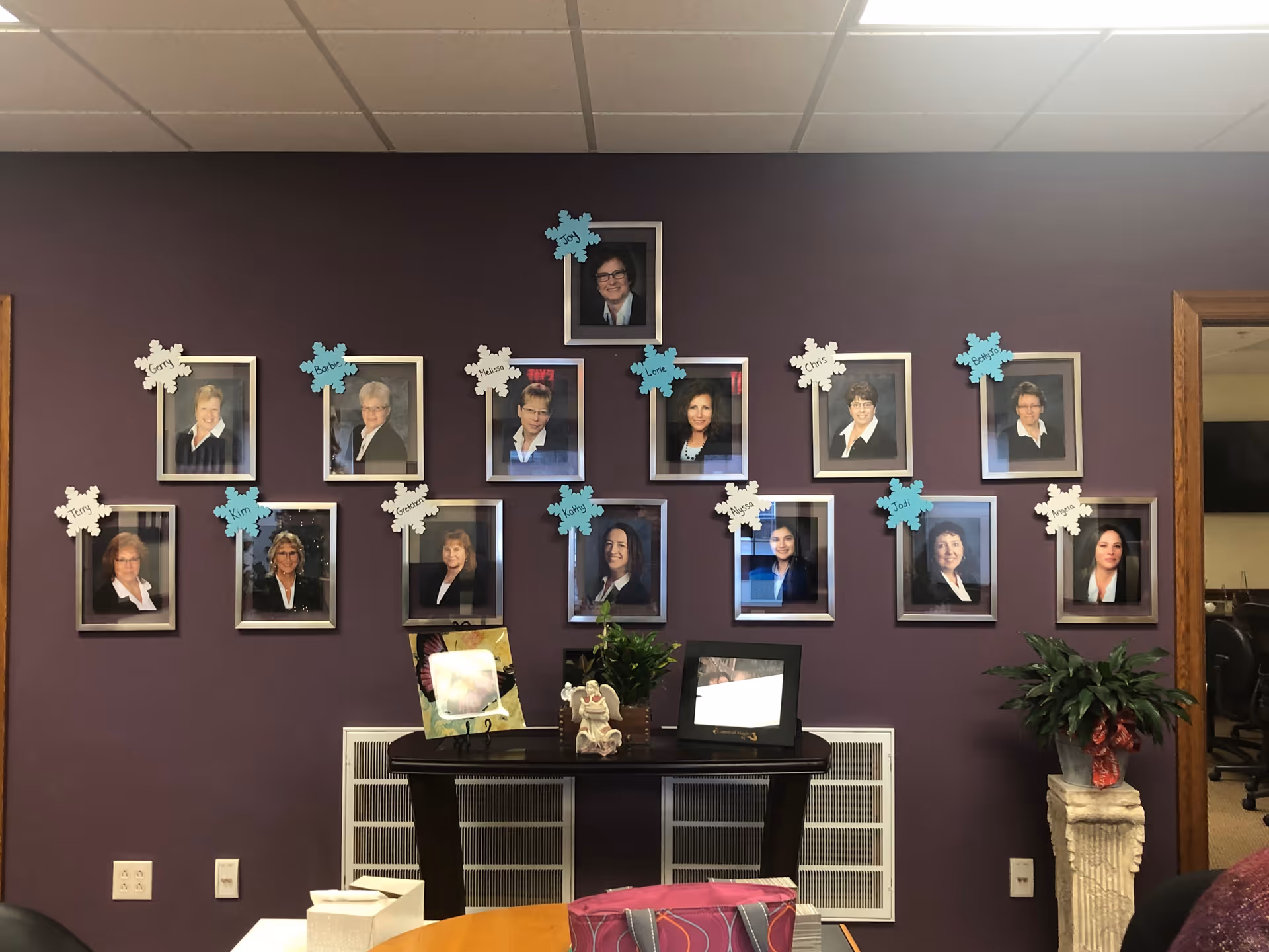 Reception area wall with a grid of framed portrait photos above a small table holding plants and picture frames.