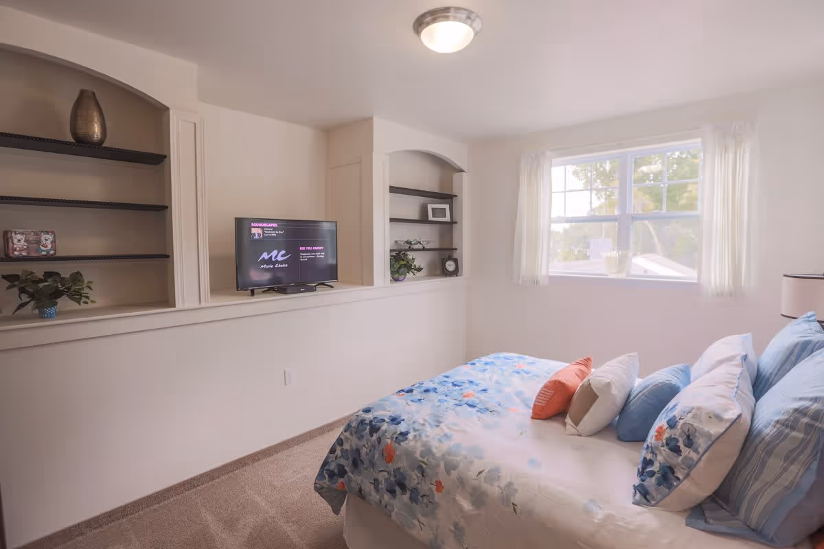 A bright bedroom with a bed covered in floral and striped pillows and a blue and white floral comforter. There is a window with sheer white curtains letting in natural light. On the opposite wall, there are built-in shelves with decorative items and a flat-screen TV.