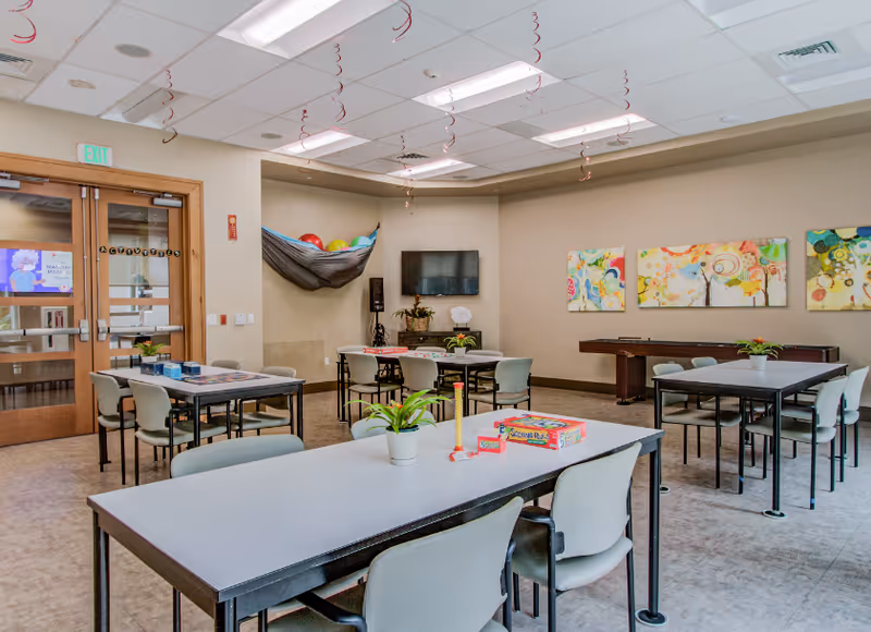 Bright communal activity room with tables and chairs, a wall-mounted TV, colorful artwork, and board games on the tables.