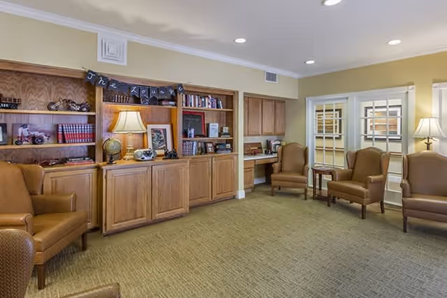 A cozy common area with beige walls and carpeted floor featuring several brown leather armchairs arranged around small wooden tables. Built-in wooden bookshelves and cabinets line one wall, decorated with books, a globe, a lamp, and framed pictures. The room is softly lit by ceiling lights and table lamps, creating a warm and inviting atmosphere.