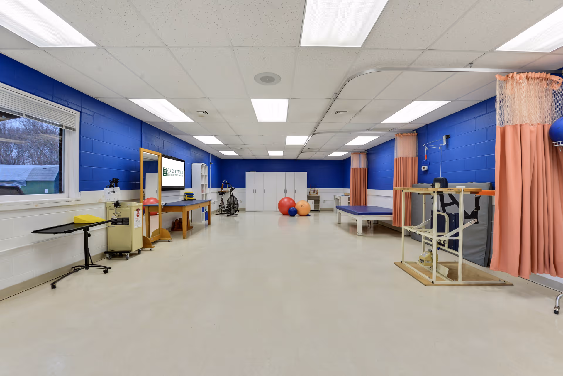 A spacious rehabilitation room with blue and white walls, fluorescent ceiling lights, and various physical therapy equipment including exercise balls, a stationary bike, therapy tables, and a wooden frame with straps. There are peach-colored privacy curtains along one wall and a window showing an outdoor view.