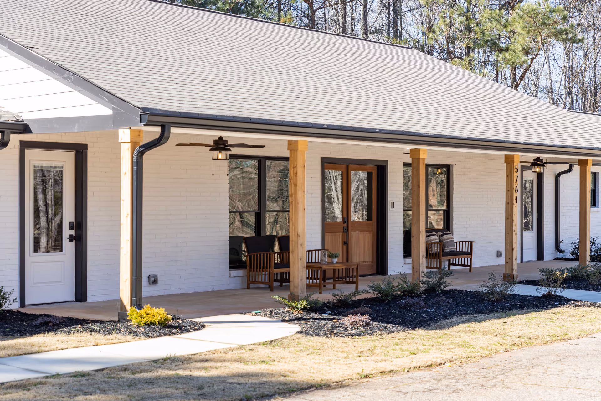 Front porch of a single-story white brick building with wooden posts, outdoor seating, and double wooden entry doors.