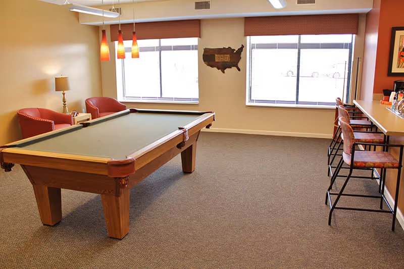 A bright common room with a pool table, bar seating, and lounge chairs under pendant lights.