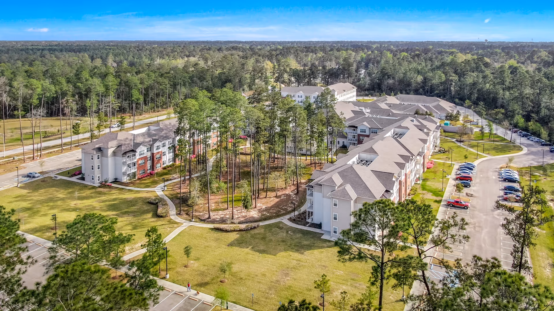 Aerial view of St. Anthony's Gardens senior living facility showing multiple connected residential buildings surrounded by tall pine trees, green lawns, walking paths, and parking lots with cars. The area is bordered by a dense forest under a clear blue sky.
