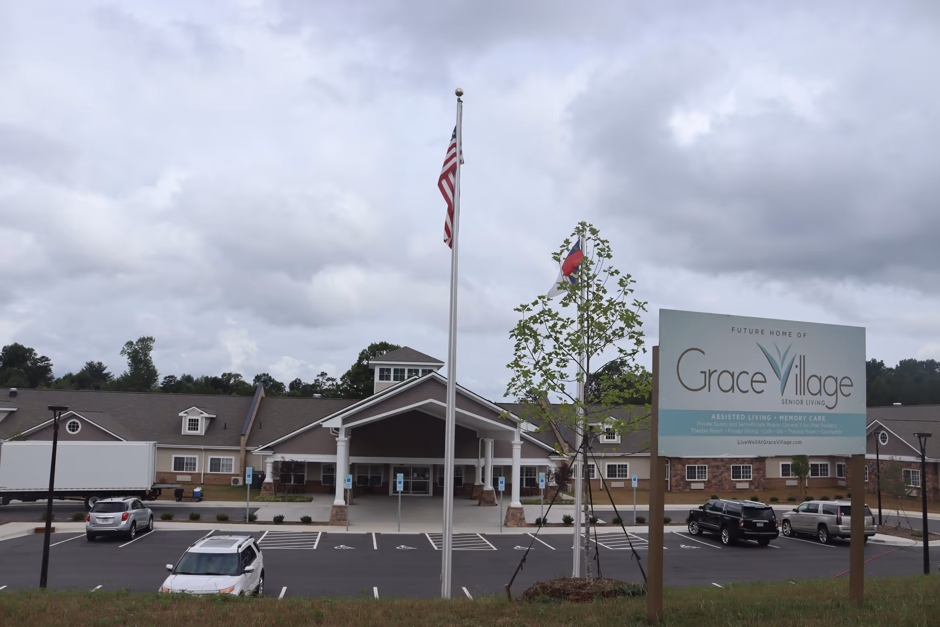 Entrance and front exterior of the Grace Village assisted living and memory care building with flagpoles, a parking lot, and a large sign.
