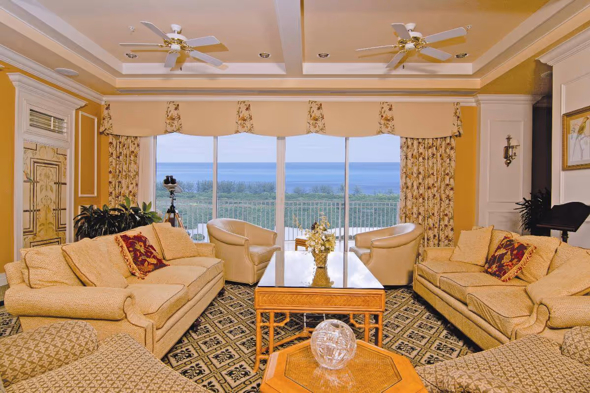 A spacious living room with two beige sofas, two beige armchairs, a glass-top coffee table with a flower arrangement, and a patterned carpet. Large windows with floral curtains offer a view of greenery and the ocean in the distance. The ceiling has two white ceiling fans and recessed lighting.
