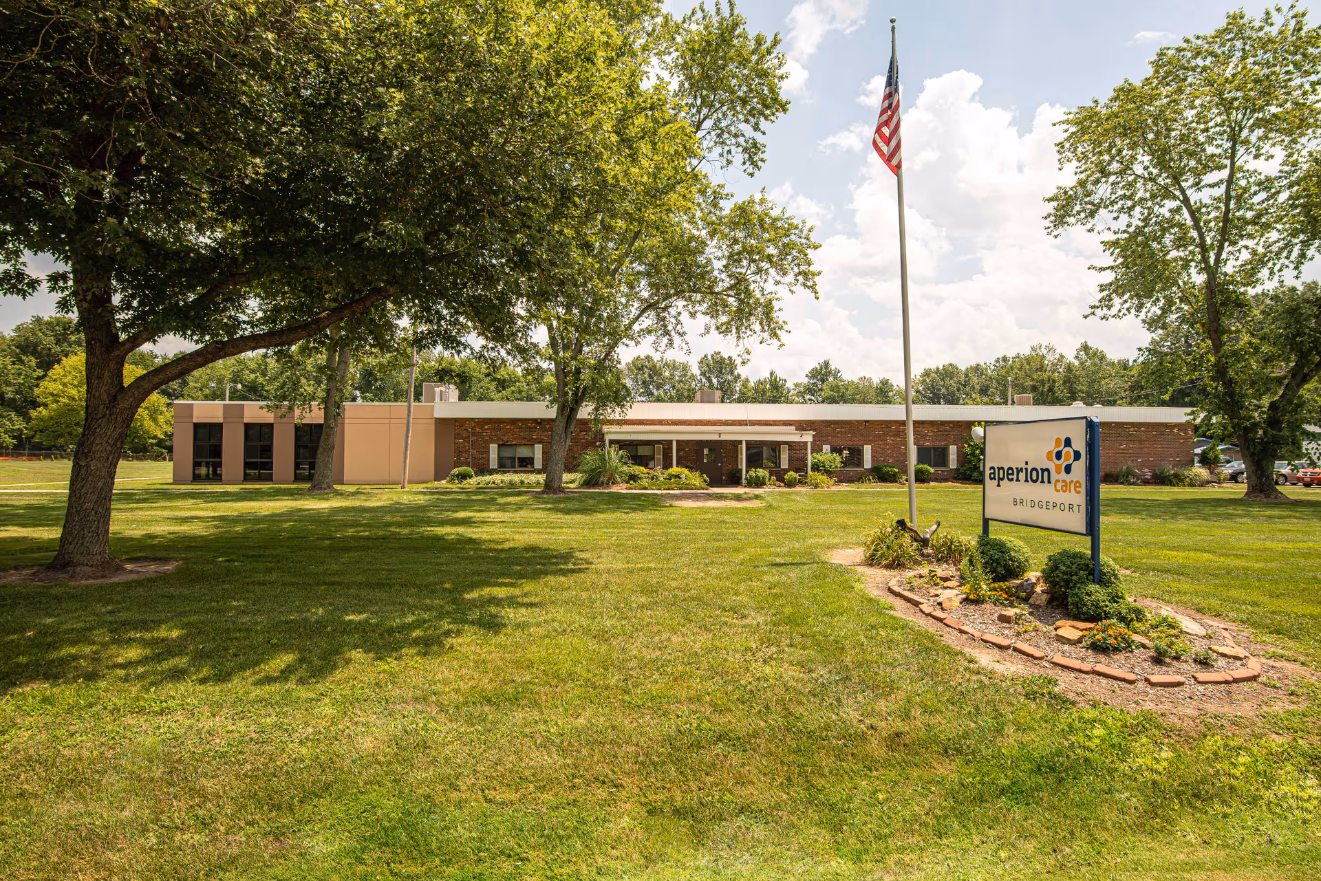 Single-story senior care building front with a large lawn, trees, an American flagpole, and a sign reading "aperion care BRIDGEPORT".