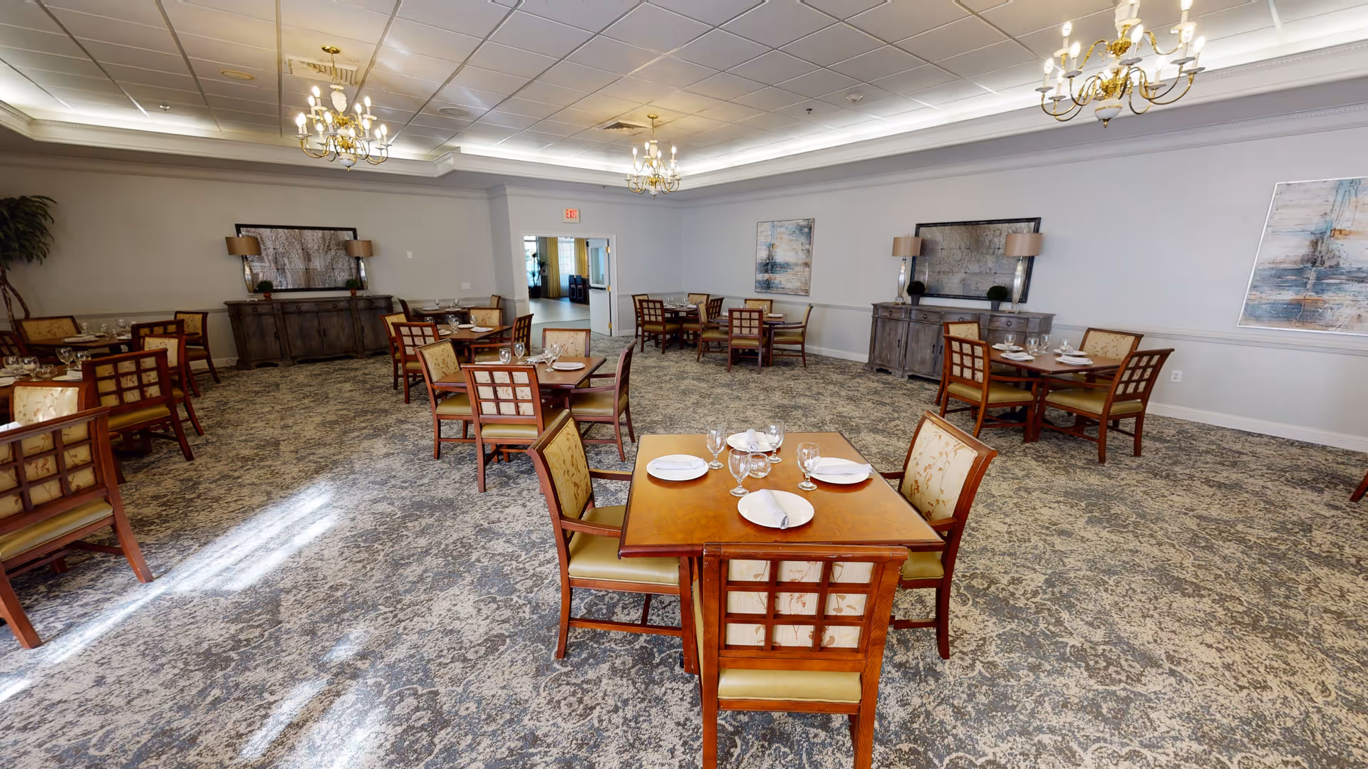 A spacious dining room with multiple wooden tables and chairs arranged neatly. Each table is set with plates, glasses, and napkins. The room features patterned carpet flooring, light gray walls adorned with abstract paintings, and two wooden sideboards with lamps. Elegant chandeliers hang from the ceiling, providing warm lighting.