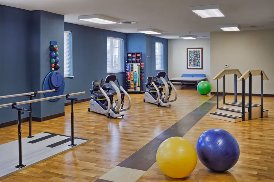 A spacious physical therapy or exercise room with wooden floors, two stationary exercise bikes, parallel bars for walking practice, colorful exercise balls, a set of stairs for rehabilitation, and shelves with various exercise equipment. The room has blue and beige walls with windows letting in natural light.