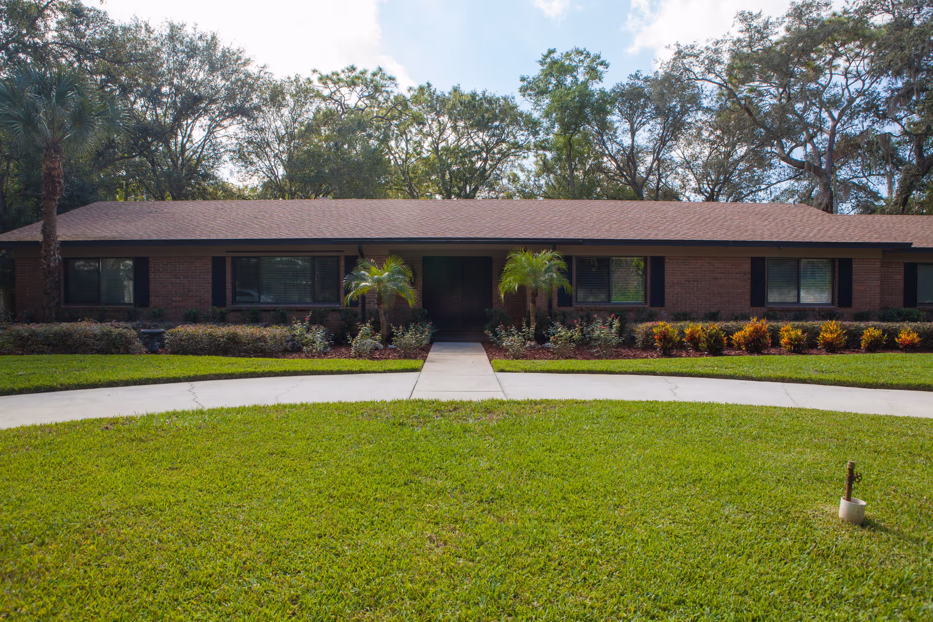 Single-story brick building with a brown shingled roof, surrounded by well-maintained green grass, shrubs, and small palm trees. A concrete walkway leads to the dark double front doors. Tall trees are visible in the background under a partly cloudy sky.