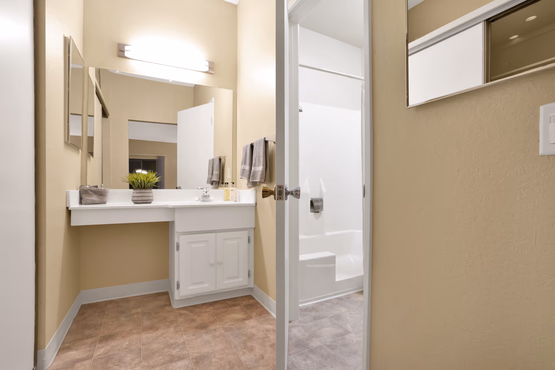 A clean bathroom with beige walls and tiled floor. There is a white vanity with a sink, a large mirror above it, and a small potted plant on the counter. Two gray hand towels hang on a towel rack next to the vanity. An open door reveals a white bathtub and shower area with a built-in seat and a towel hanging on the shower wall.