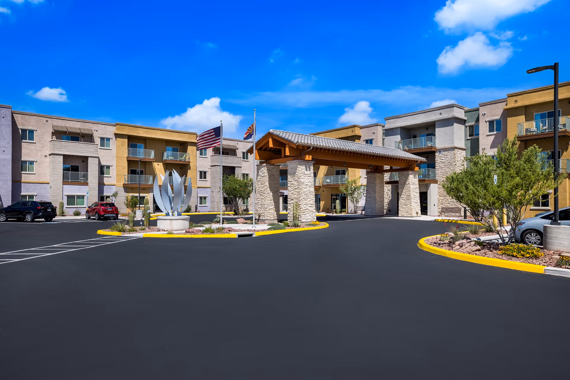 Exterior view of a senior living facility with a large covered entrance supported by stone pillars, multiple three-story buildings with balconies, a sculpture, and two flagpoles with American and state flags under a bright blue sky.