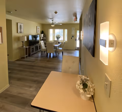 Interior view of a living and dining area in an assisted living facility. The space features a small table with a white flower arrangement in the foreground, a dining table with chairs in the middle, and a TV on a stand against the wall. The room has light-colored walls, wood flooring, and a ceiling fan. Natural light comes through a window and a glass door at the far end.