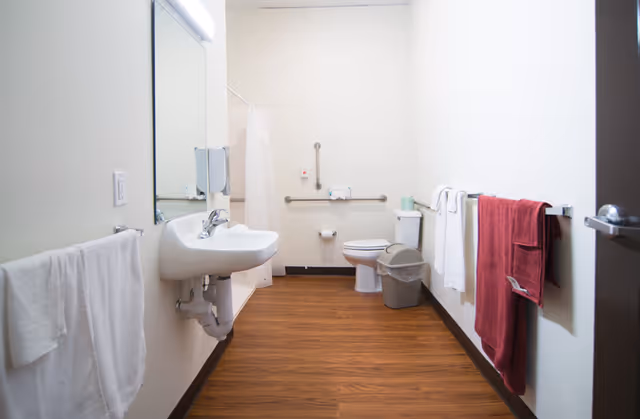 A clean and accessible bathroom with a white sink, mirror, toilet, and shower area. The room has wooden flooring and white walls. There are grab bars near the toilet and a trash can beside it. Several towels hang on racks along the walls.