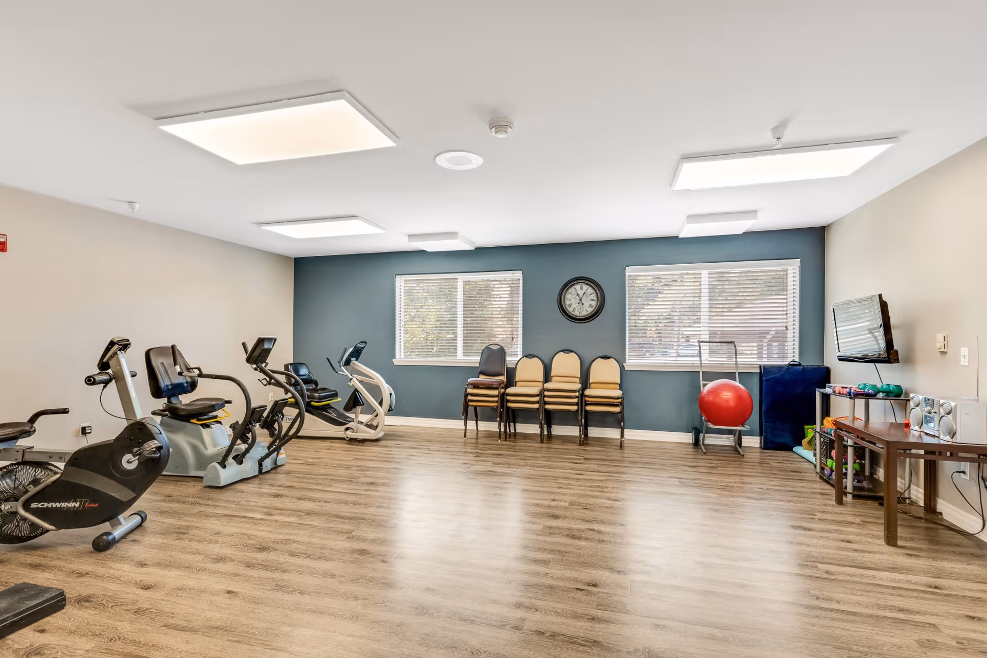 Bright exercise/activity room with stationary bikes, stacked chairs under a wall clock, a TV, and additional fitness equipment on wood floors.