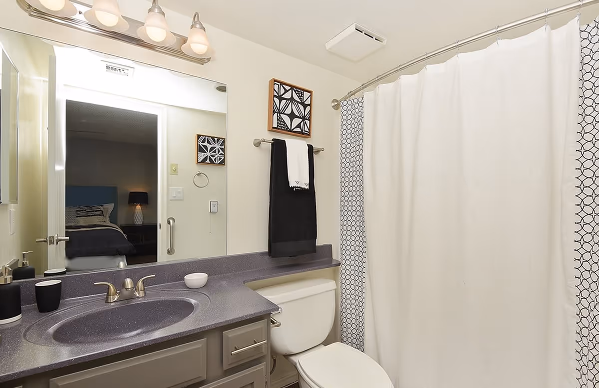 A clean and modern bathroom featuring a gray countertop with an integrated sink, a toilet, and a shower with a white curtain and patterned liner. Above the sink is a large mirror reflecting a bedroom with a bed and lamp. A towel rack holds a black towel with a white towel on top, and a small framed black and white geometric artwork hangs on the wall.