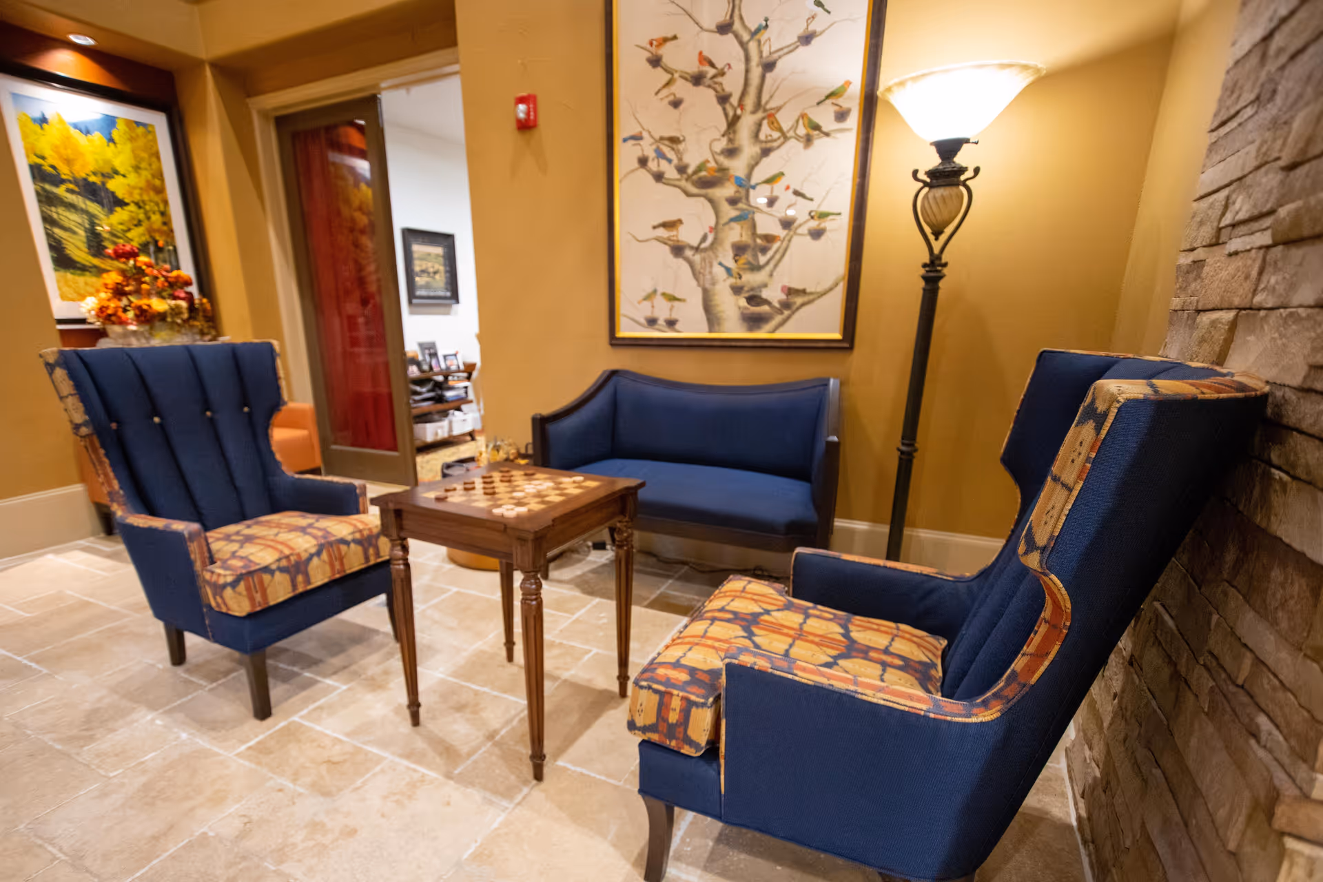 Cozy seating area with two patterned armchairs, a small game table, a blue loveseat, a floor lamp, and bird artwork on the wall.