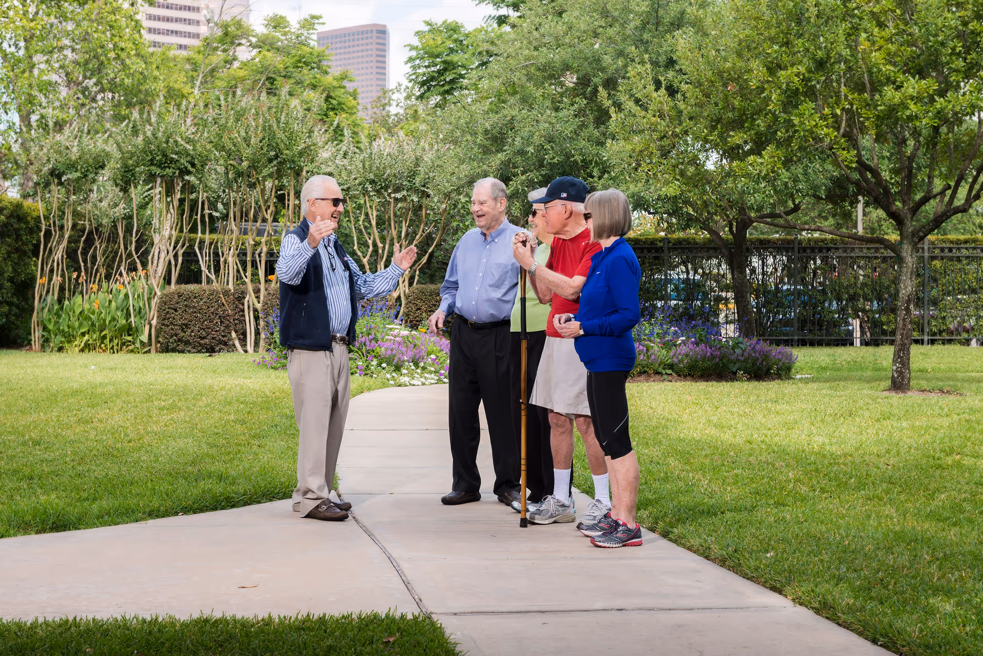 Four elderly people standing and talking on a paved pathway in a green garden area with trees, bushes, and flowers. One man is gesturing with his hands while the others listen and smile. The background shows some tall buildings partially visible behind the trees.
