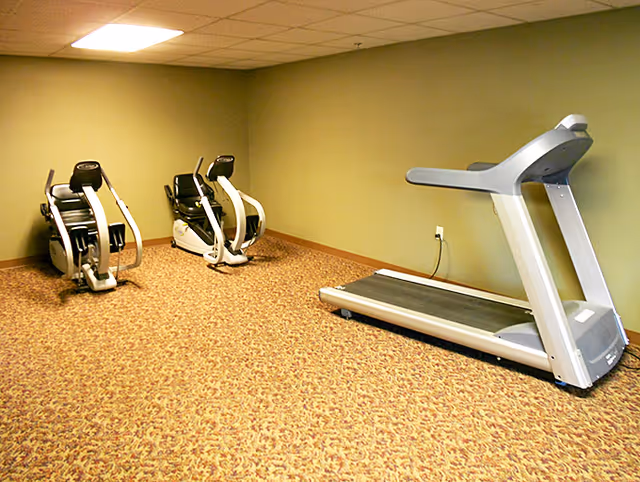 A small exercise room with two recumbent stationary bikes and one treadmill. The room has beige patterned carpet, light green walls, and a drop ceiling with fluorescent lighting.