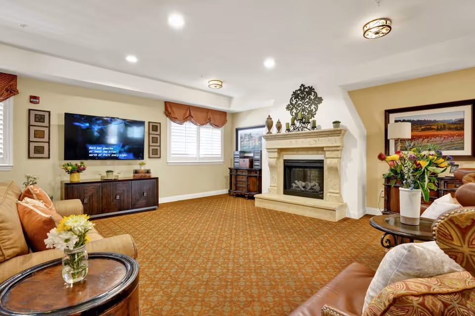 A cozy living room with a patterned carpet, beige walls, and a stone fireplace. The room features a large flat-screen TV mounted on the wall above a wooden cabinet, several comfortable sofas and armchairs with cushions, and side tables with flower vases. There are two windows with white blinds and brown valances, and framed artwork on the walls.
