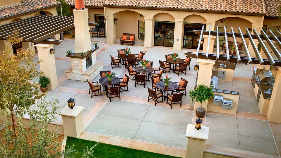 Outdoor patio area with multiple round tables surrounded by chairs, each table decorated with a potted plant. The space features a pergola, a built-in grill, and a fireplace, with beige walls and tiled flooring. There are also some potted plants and greenery around the patio.