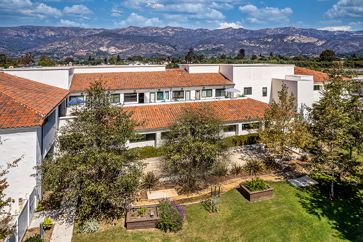 Mediterranean-style senior living building with red tile roofs surrounding a landscaped courtyard and mountains beyond.