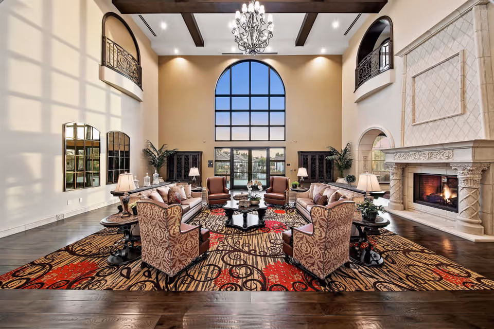 Spacious and elegant senior living facility common area with high ceilings, large arched window, chandelier, two sofas, four armchairs, a patterned area rug, side tables with lamps, decorative plants, and a large stone fireplace with a fire burning.