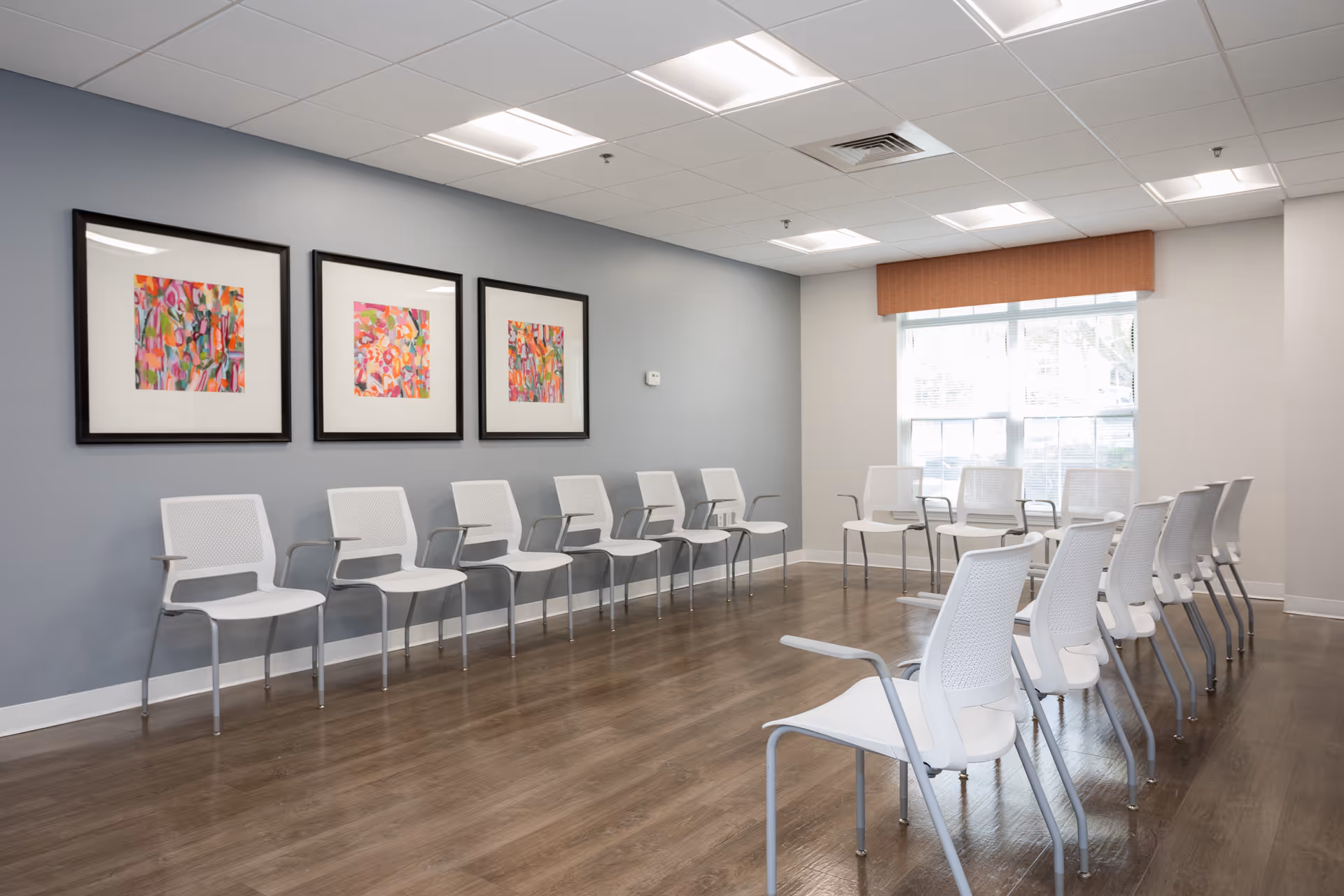 A bright room with white chairs arranged in a semi-circle along the walls and in rows. The room has wooden flooring, a gray accent wall with three colorful abstract paintings, and a large window with blinds and an orange valance allowing natural light to enter.