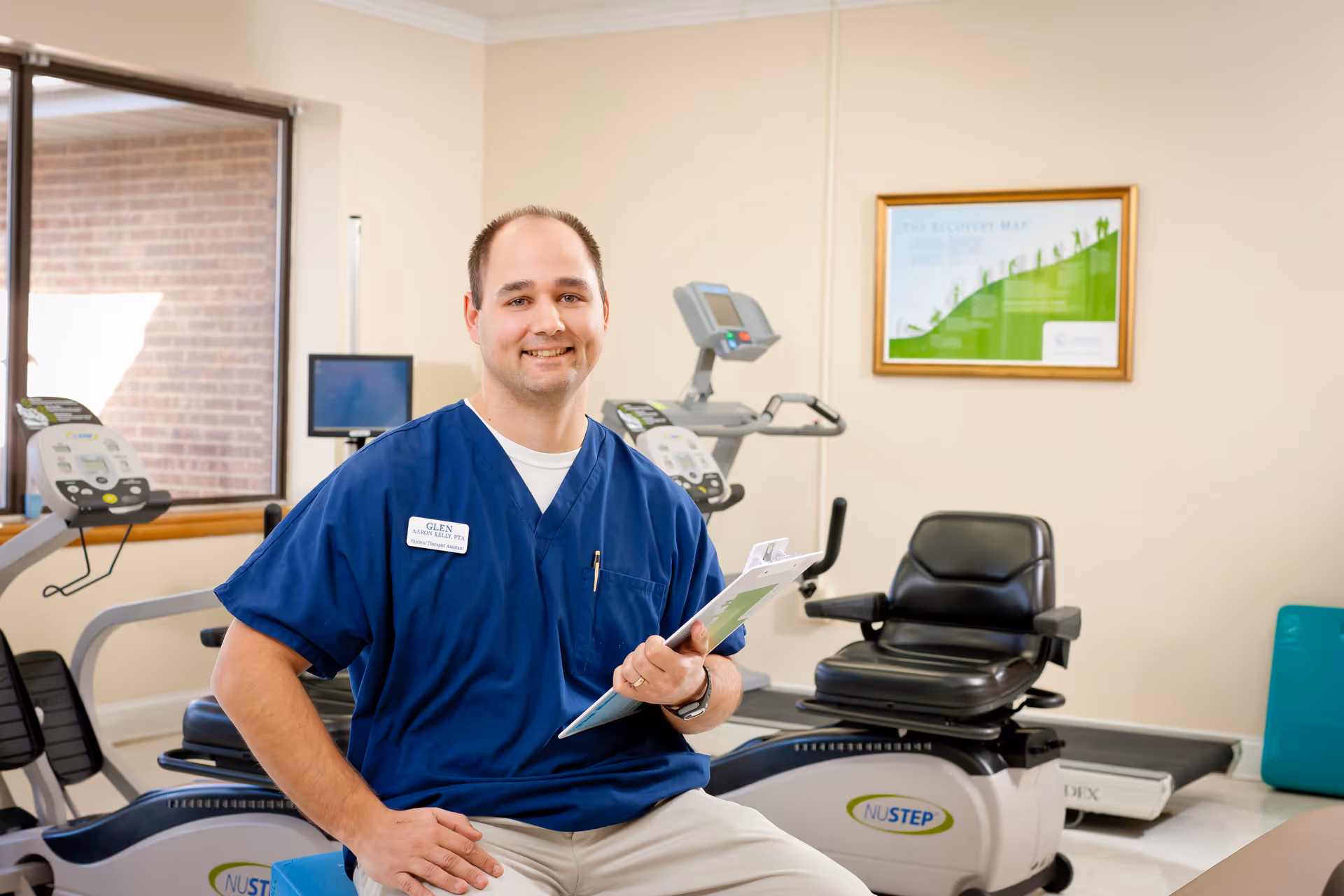 A man in blue scrubs with a name tag that reads Glen is sitting on a blue exercise ball in a fitness room. Behind him are exercise machines including a NuStep recumbent cross trainer. The room has beige walls, a window with a view of a brick wall, and a framed chart on the wall.
