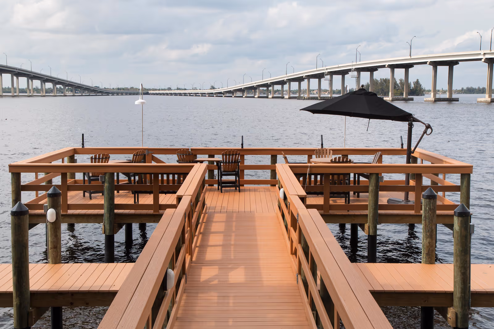 A wooden dock extending over a body of water with several Adirondack chairs and a table with a large black umbrella. In the background, there is a long bridge spanning across the water under a cloudy sky.