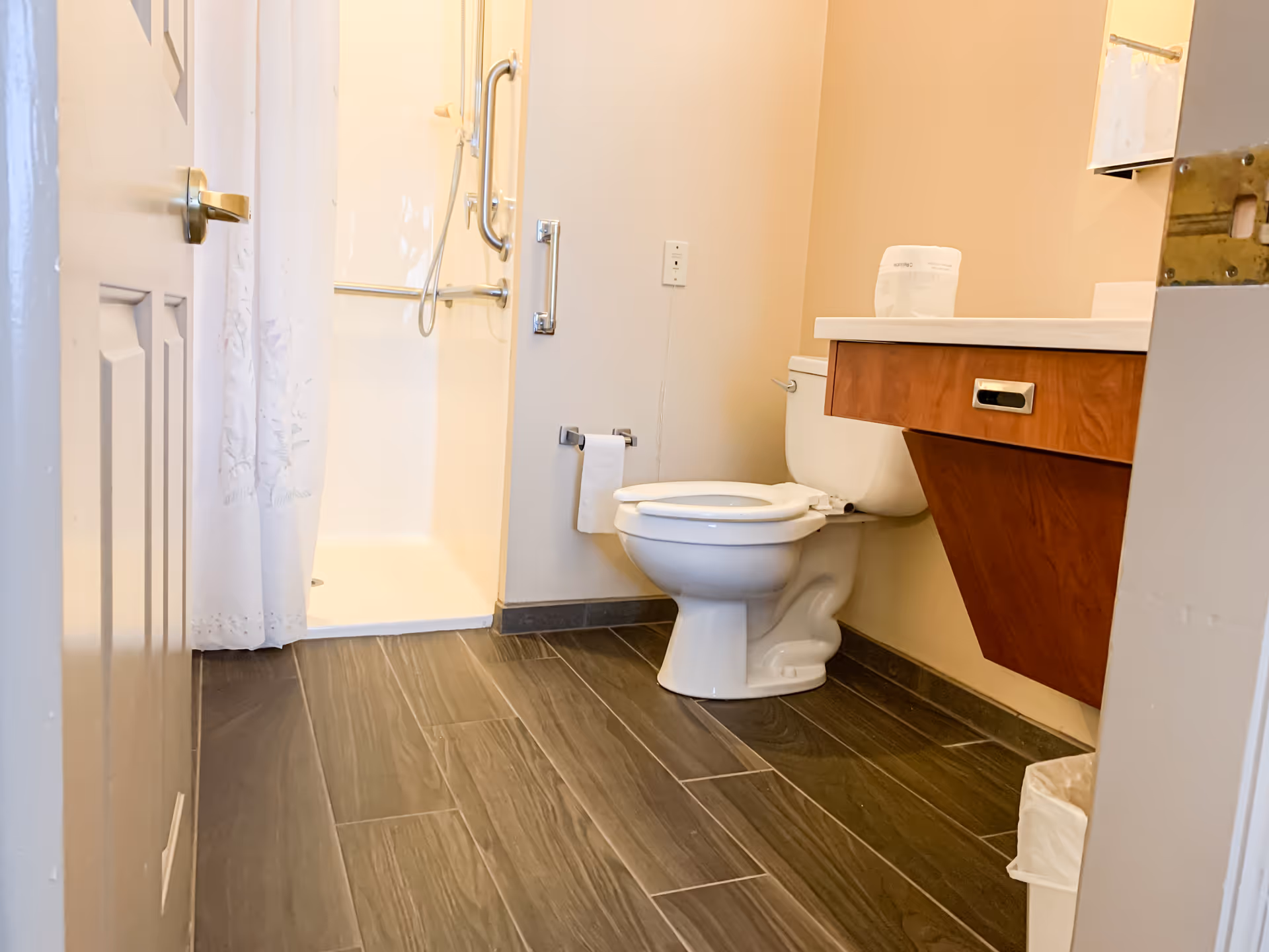 A clean bathroom with a white toilet, a wooden vanity with a white countertop, a shower with a white curtain, and gray tiled floor.