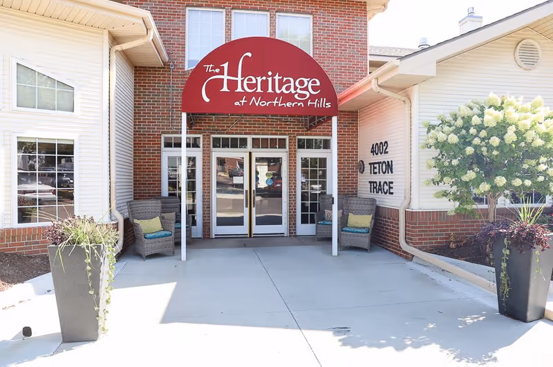 Entrance to The Heritage at Northern Hills facility with a red awning displaying the facility name above double glass doors. The building exterior features red brick and white siding, with two wicker chairs with cushions on either side of the entrance and large planters with greenery.