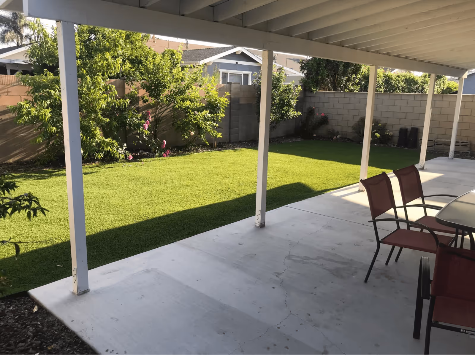 Covered patio area with concrete flooring and red chairs around a glass table, overlooking a green lawn and garden with trees and bushes enclosed by a brick wall.