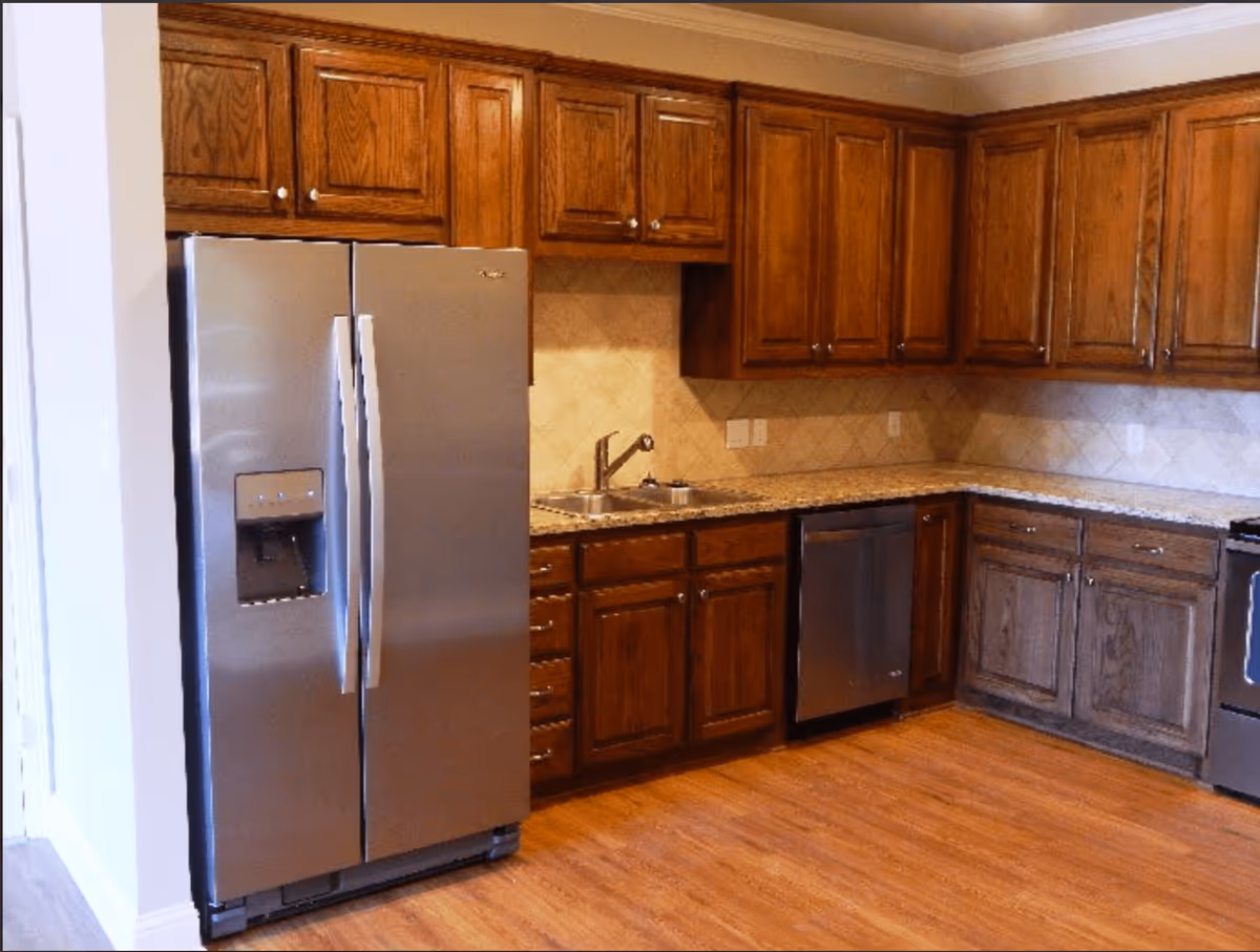 A kitchen with wooden cabinets, a stainless steel refrigerator with a water and ice dispenser, a double sink, a dishwasher, and a stove. The floor is wood, and the backsplash has a beige tile pattern.