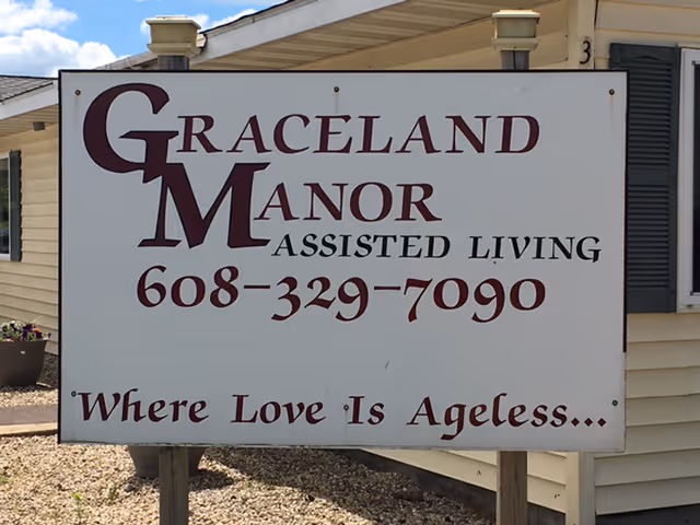 A large white sign outside a beige building with dark shutters that reads 'Graceland Manor Assisted Living 608-329-7090 Where Love Is Ageless...'. The sign is mounted on wooden posts and there is gravel on the ground around it.