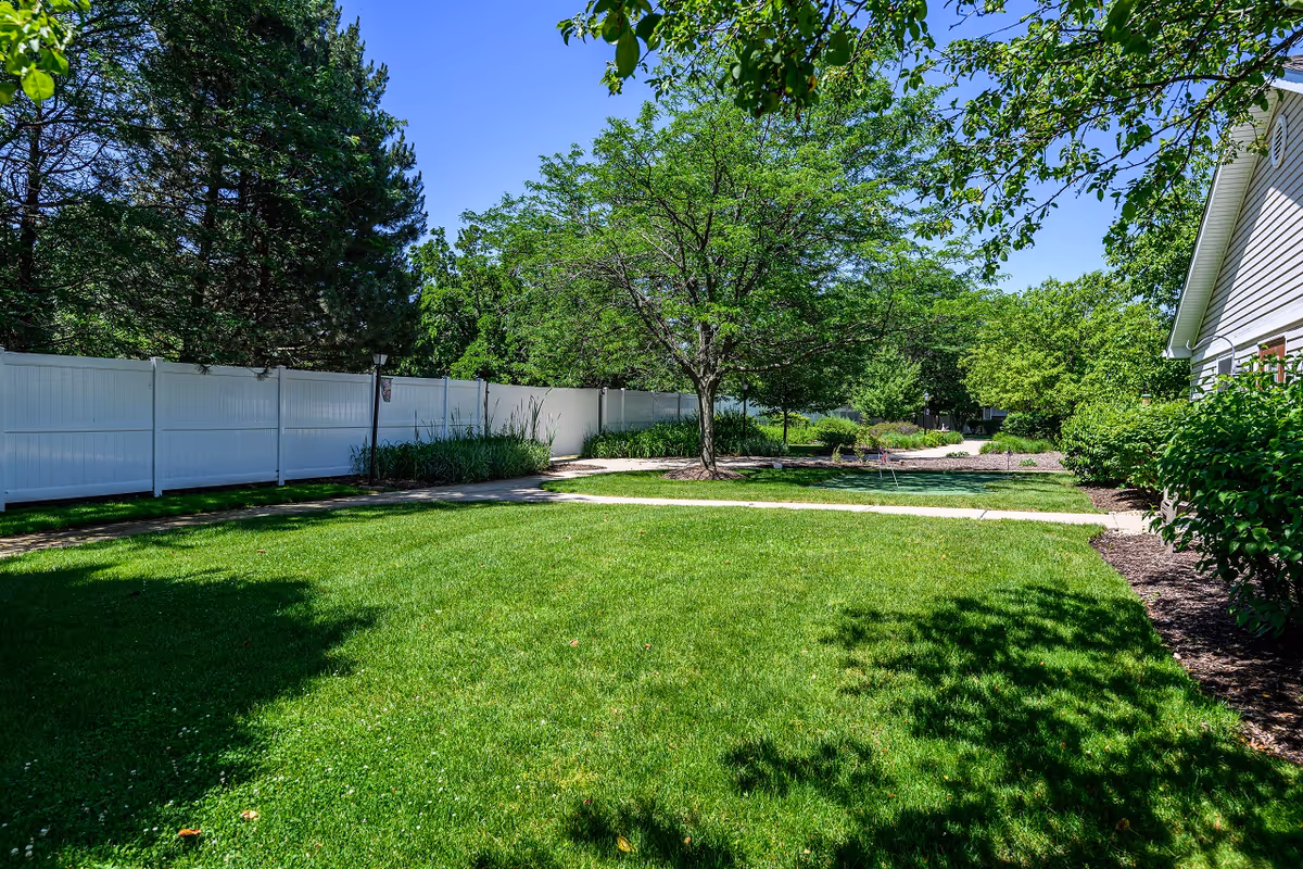 A sunny outdoor garden area with a well-maintained green lawn, a paved walkway, several trees, bushes, and a white fence along the left side. Part of a building with beige siding is visible on the right side.