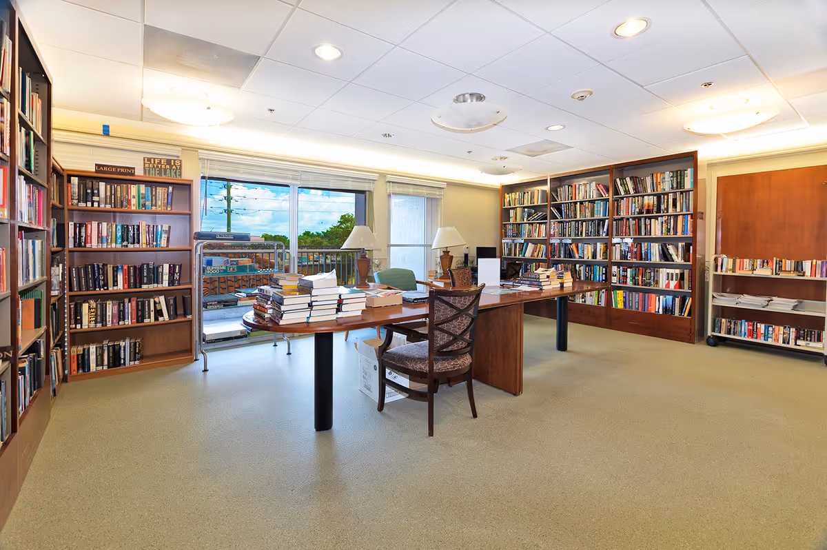 A bright senior living library with bookshelves along the walls, a central table stacked with books, chairs, lamps, and large windows overlooking the outdoors.