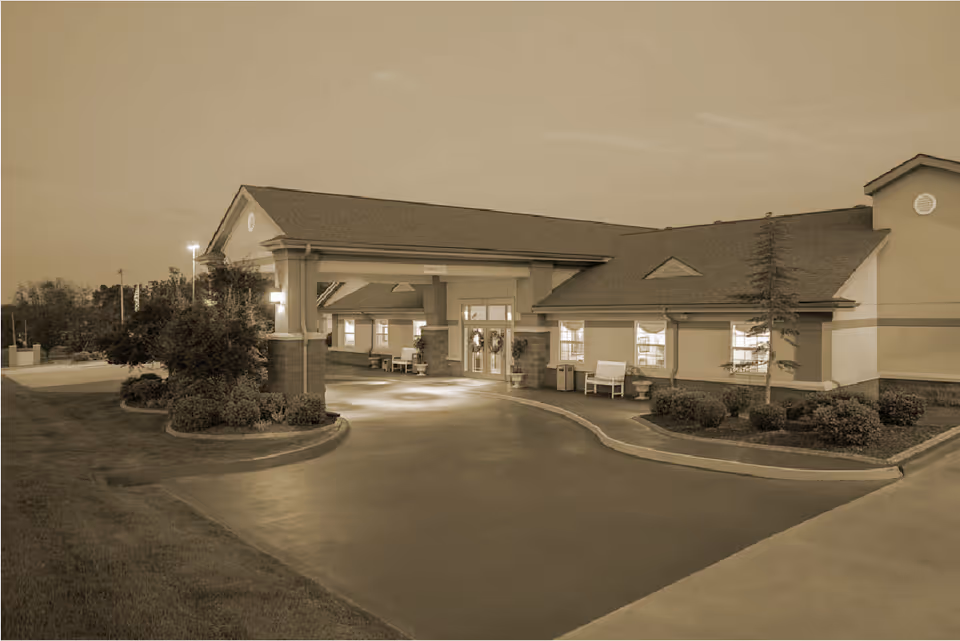 Exterior view of the entrance to a nursing and rehabilitation facility at dusk, featuring a covered drop-off area, benches, landscaped bushes, and a paved driveway.