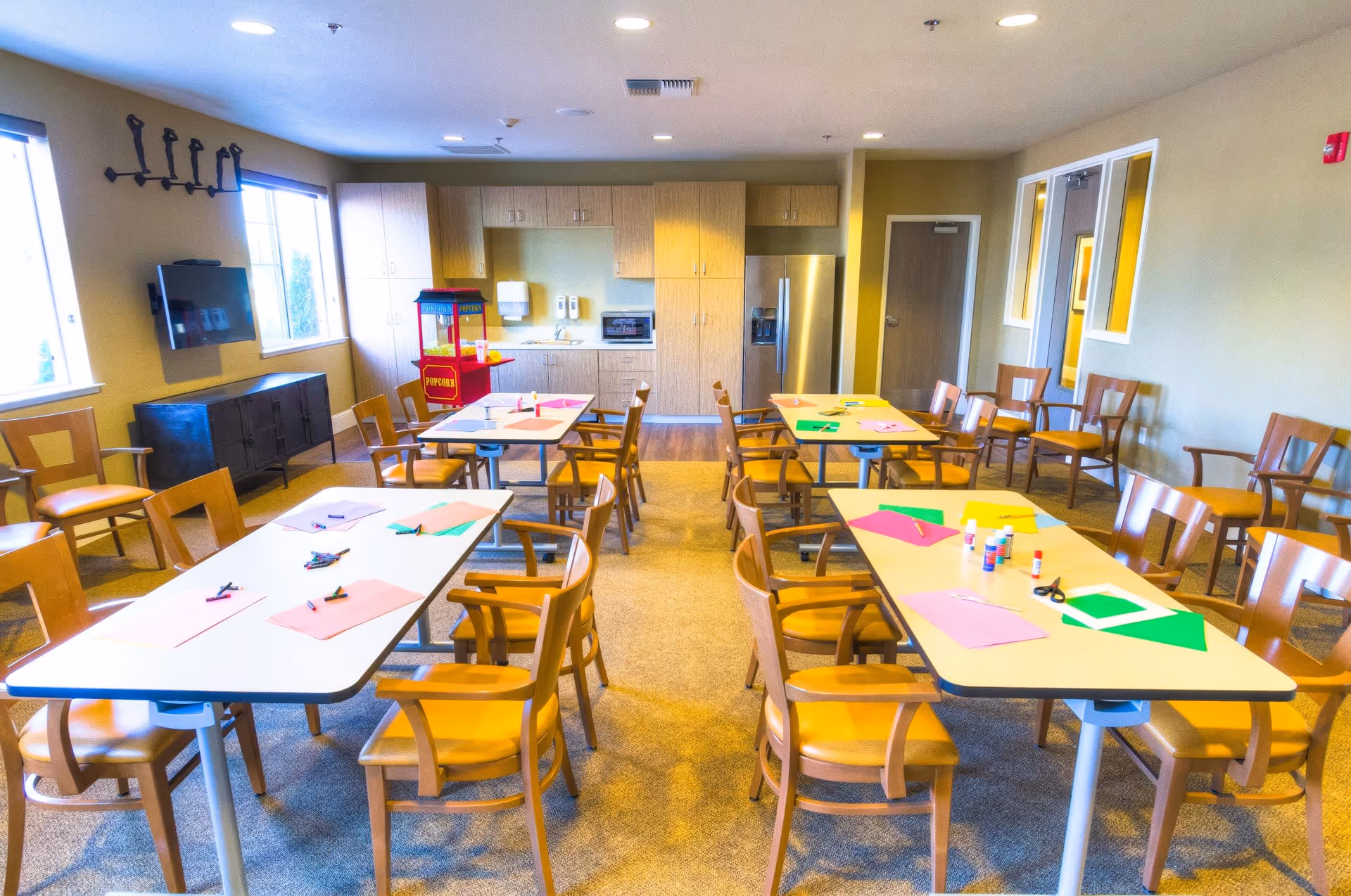 A bright activity room with several tables and wooden chairs arranged around them. The tables have colorful craft supplies including paper, glue sticks, scissors, and crayons. The room has large windows letting in natural light, a popcorn machine, a wall-mounted TV, and a kitchenette area with cabinets, a microwave, and a refrigerator.