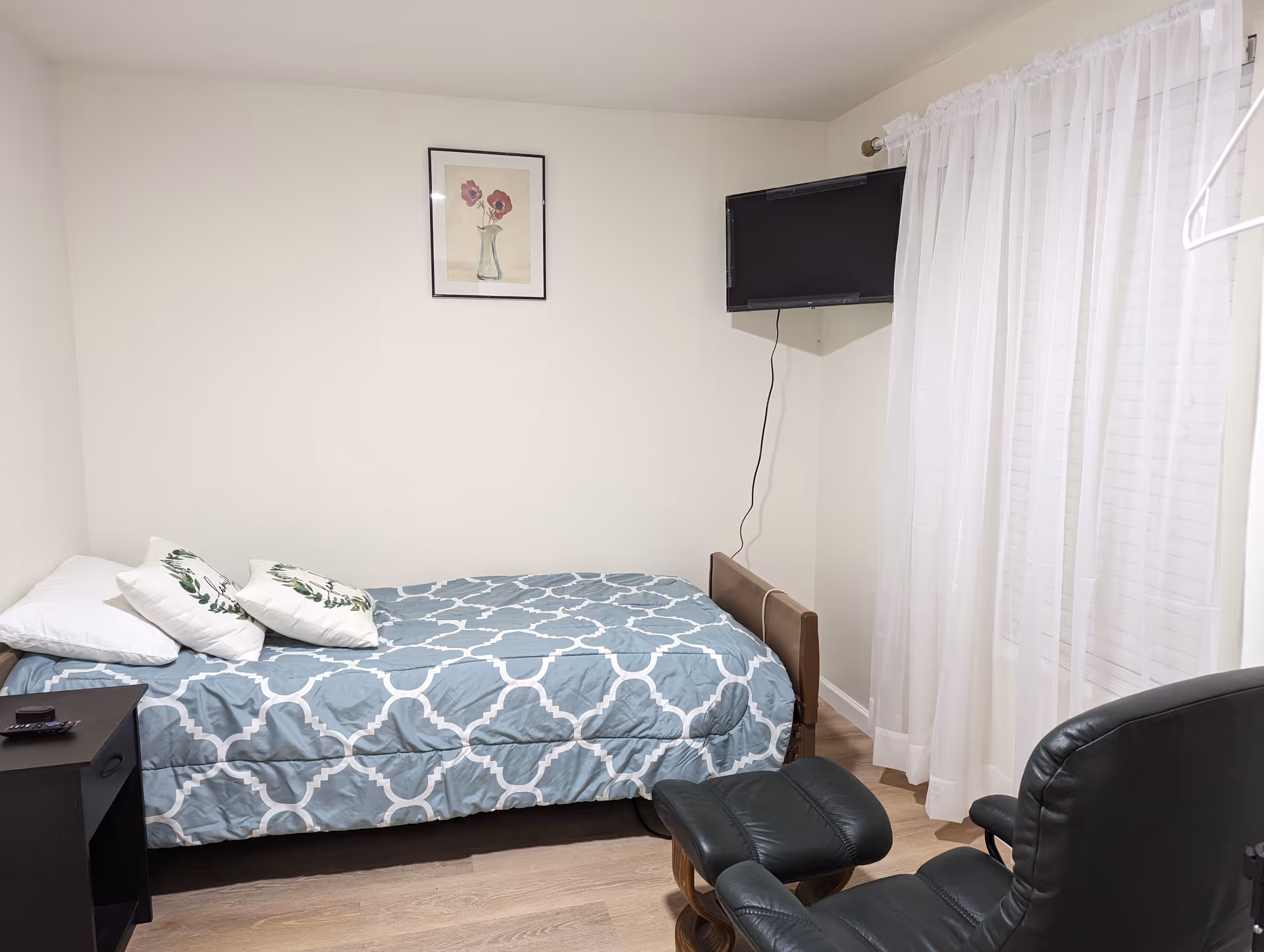 A simple bedroom with a twin bed in blue patterned bedding, a wall-mounted TV, a reclining chair, nightstand and sheer curtains over a window.
