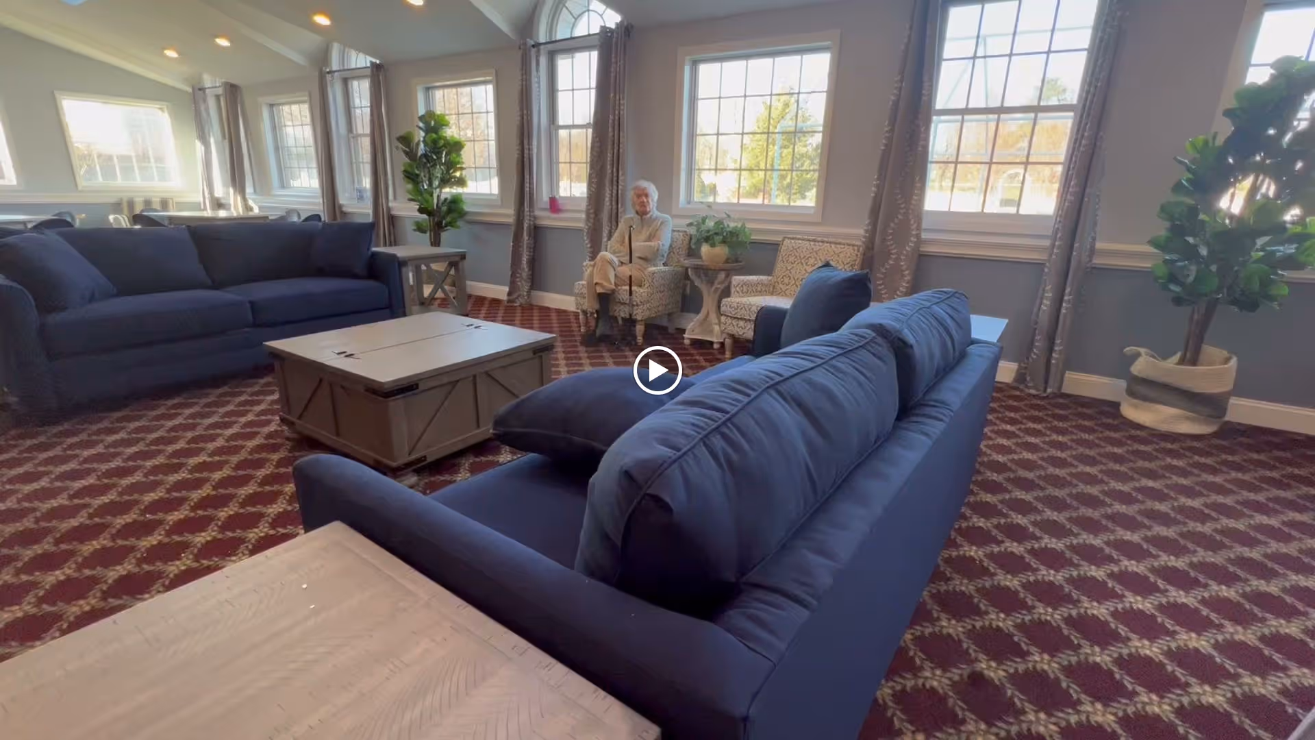 A sunny communal lounge with navy sofas, a central coffee table, patterned carpet, large windows and a person seated by the window.