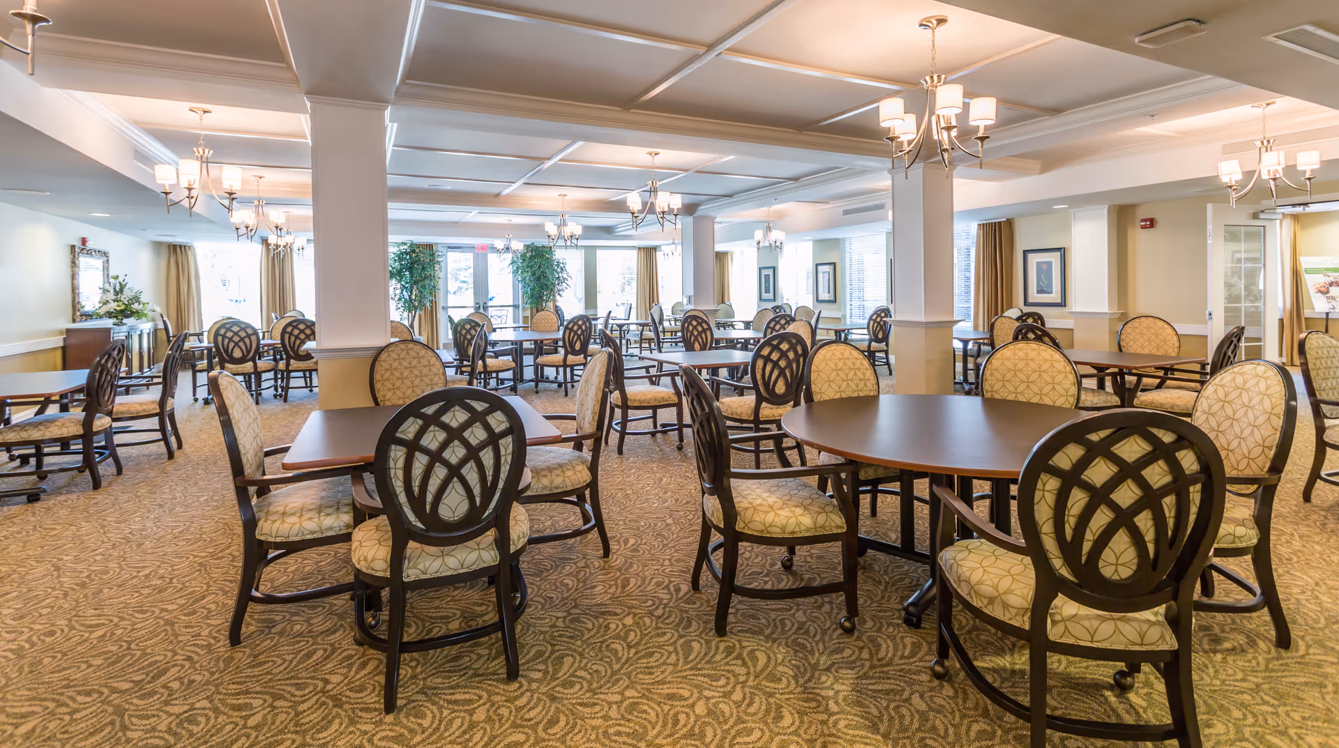 A spacious dining room with multiple round and rectangular tables surrounded by cushioned chairs with patterned upholstery. The room features carpeted floors, white coffered ceilings with chandeliers, large windows with curtains, and decorative plants near the windows.